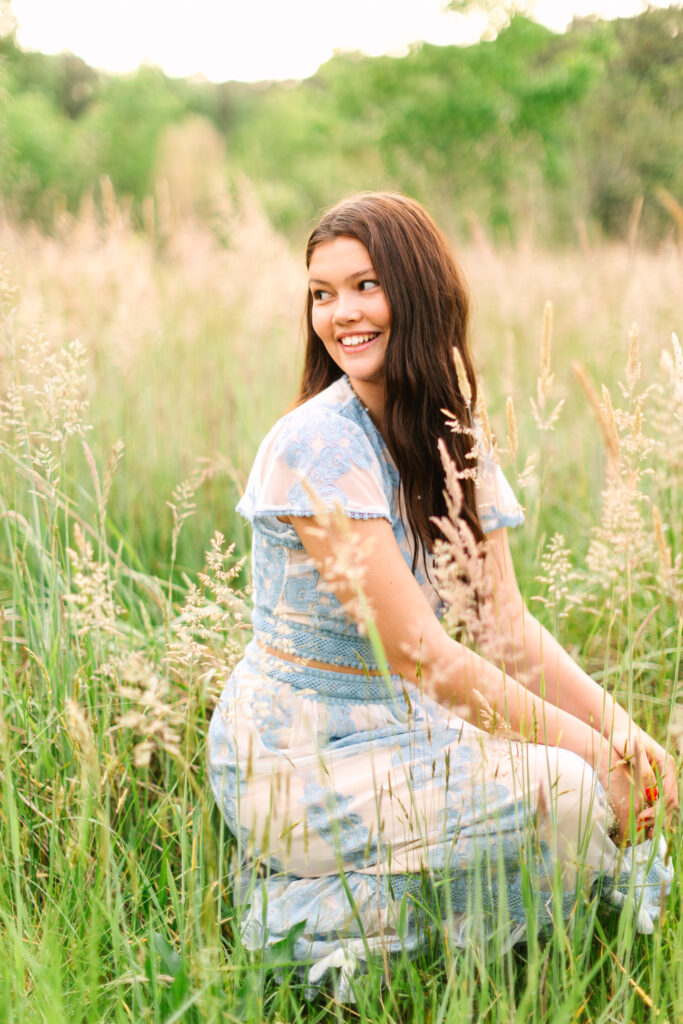 Senior laughing in a sunlit field with tall grass, wearing a blue dress and surrounded by warm evening light.