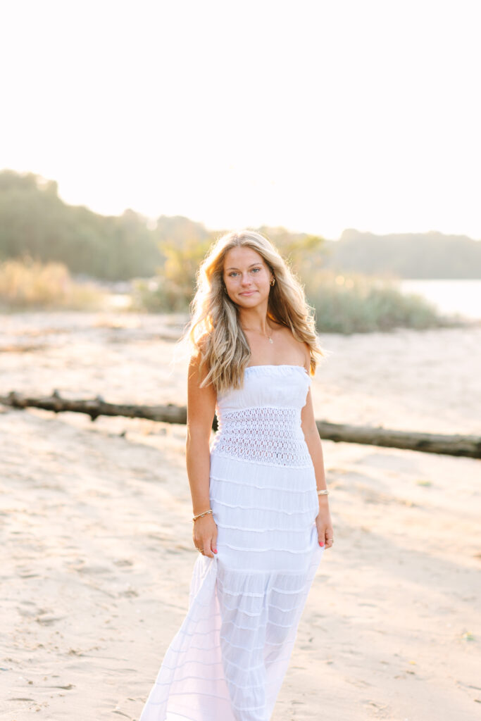 Senior girl walking along Yorktown Beach at golden hour, wearing a white dress with soft, glowing light and a calm, natural backdrop.