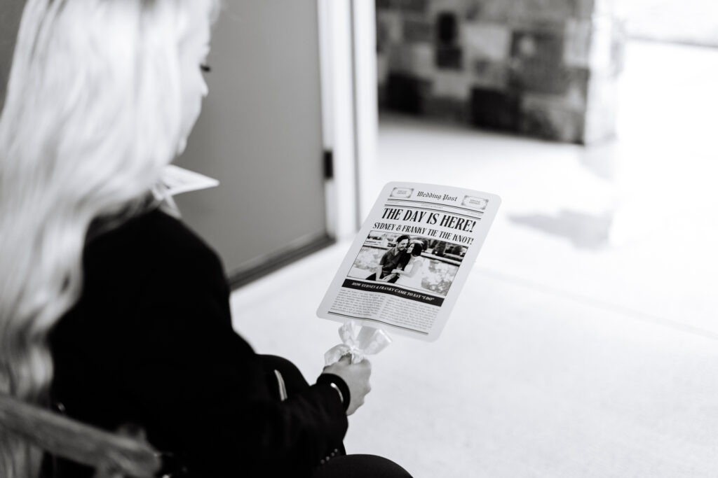 Guest holding a “The Day Is Here” newspaper-style wedding fan at The Maine of Williamsburg