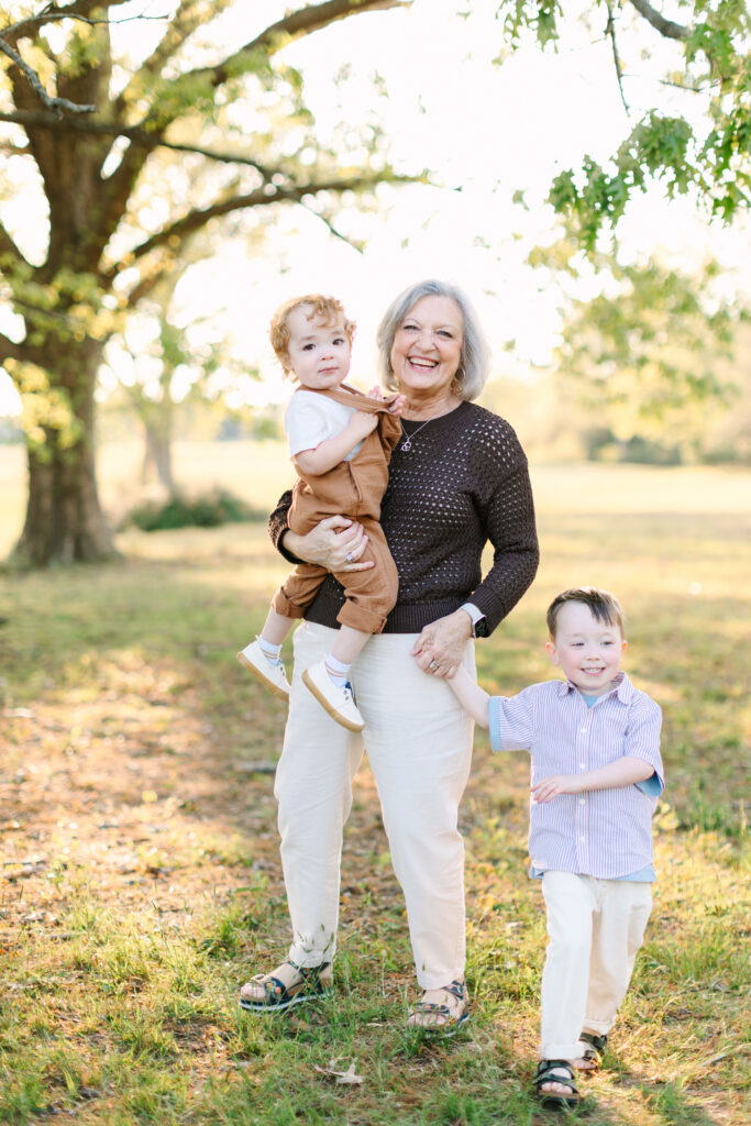 Grandmother holding one grandson and walking hand in hand with another through a field at sunset.